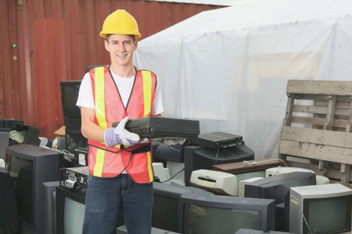 A young male worker wearing a yellow safety helmet, a high-visibility vest with red and yellow reflective stripes, and white work gloves, standing outdoors on a paved area. He is smiling while holding a black electronic device, possibly a remote control or a power tool. Behind him, there are several discarded, old electronic appliances such as CRT monitors, printers, and other obsolete computer equipment arranged on the ground. The background features a wooden fence, a large white temporary structure or tent, and a wooden pallet leaning against the fence. The scene appears set in a waste management or rubbish removal environment, with the focus on clearing or recycling electronic waste, aligning with services similar to those provided by House Clearance Addlestone in the local area.