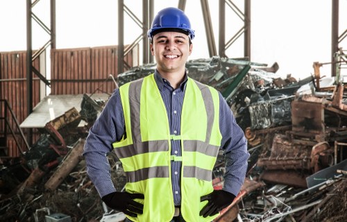 A young male worker wearing a blue safety helmet and a high-visibility yellow safety vest over a dark blue shirt stands indoors in front of a pile of assorted rubbish and debris. The rubbish includes broken wooden planks, metal scrap, and other discarded materials, indicating a waste clearance or rubbish removal scene. The background features a partially open industrial structure with metal framing and large windows allowing natural light to illuminate the workspace. The worker is smiling confidently with hands on hips, embodying a professional approach to rubbish removal services typically associated with companies like House Clearance Addlestone operating in the Addlestone area postcode. This image visually represents the transportation or clearing of waste from a property or site, highlighting safety gear and the array of materials involved in rubbish disposal tasks.