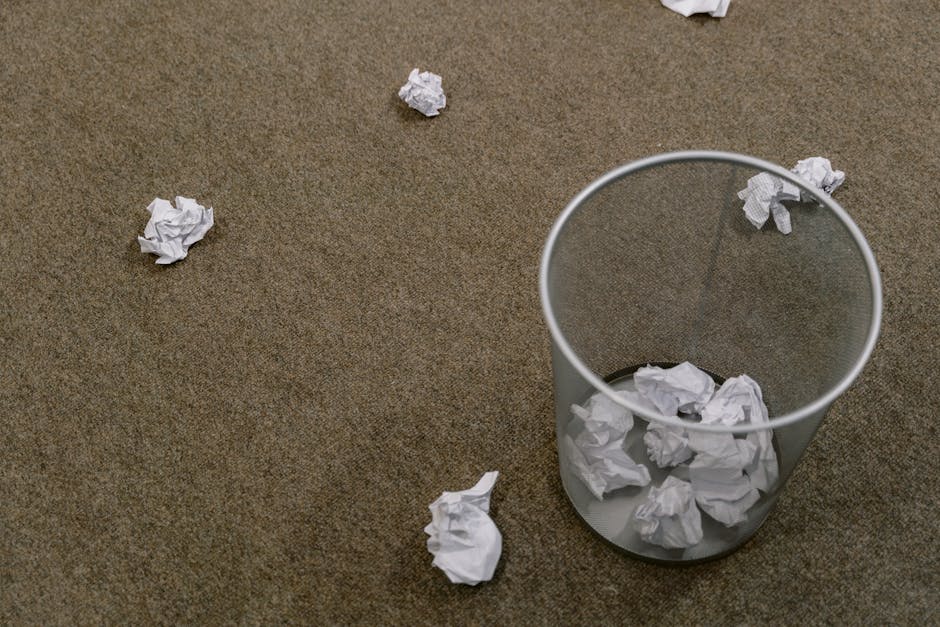 A transparent cylindrical waste bin made of clear plastic is positioned on a brown, textured carpeted floor in an indoor setting. The bin contains several crumpled sheets of白 paper, and additional c