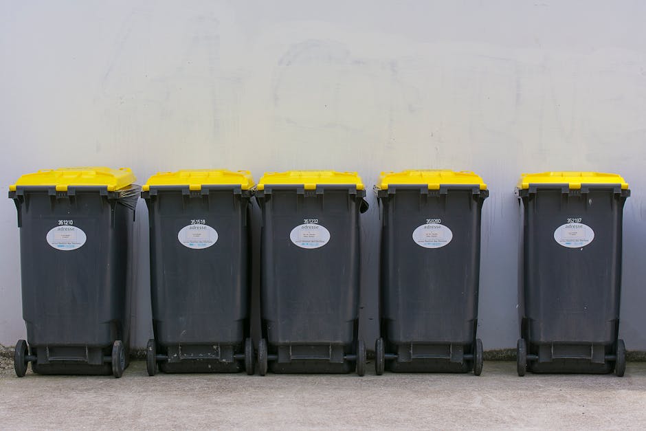 A transparent cylindrical waste bin made of clear plastic is positioned on a brown, textured carpeted floor in an indoor setting. The bin contains several crumpled sheets of白 paper, and additional c
